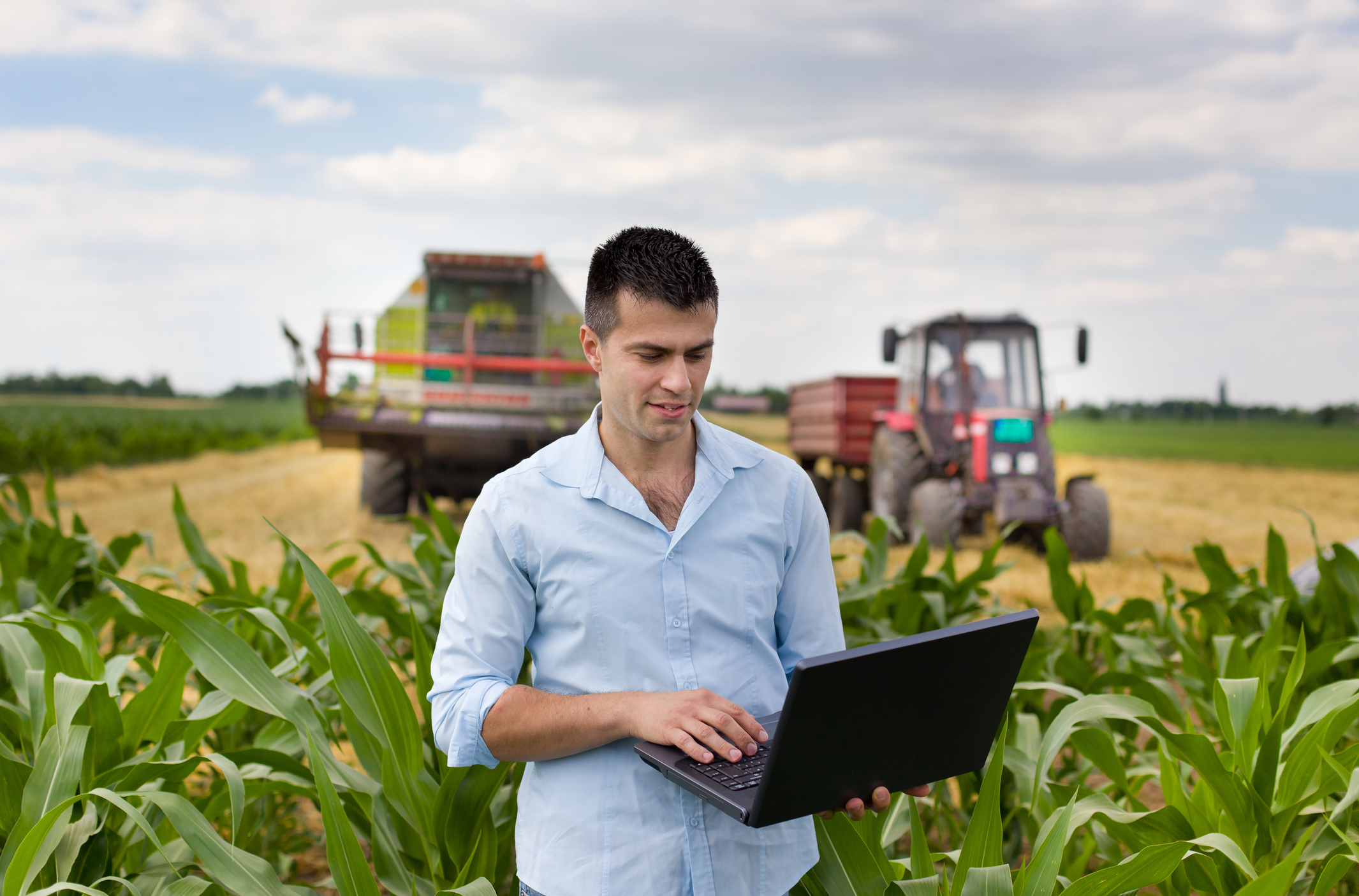 Farmer with laptop during harvest Young attractive farmer with laptop standing in corn field, tractor and combine harvester working in wheat field in background